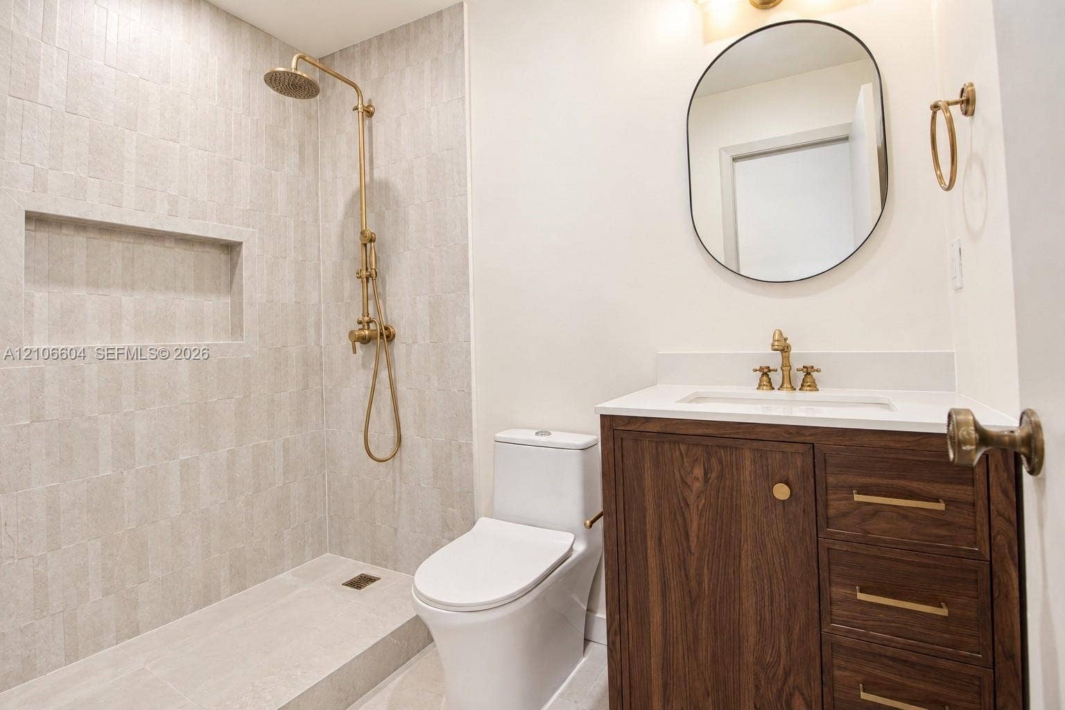 Bathroom featuring a wood-finish vanity with a stone countertop, wall-mounted oval mirror, brass-toned fixtures, walk-in shower with vertical tile surround, and a built-in shower niche