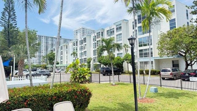 17570 Atlantic Boulevard, #105, Sunny Isles Beach, Sunny Isles Beach, FL 33160 Multi-story residential building featuring a white facade, uniform multi-pane windows, black metal perimeter fencing, manicured landscaping with mature palms, and a dedicated parking area