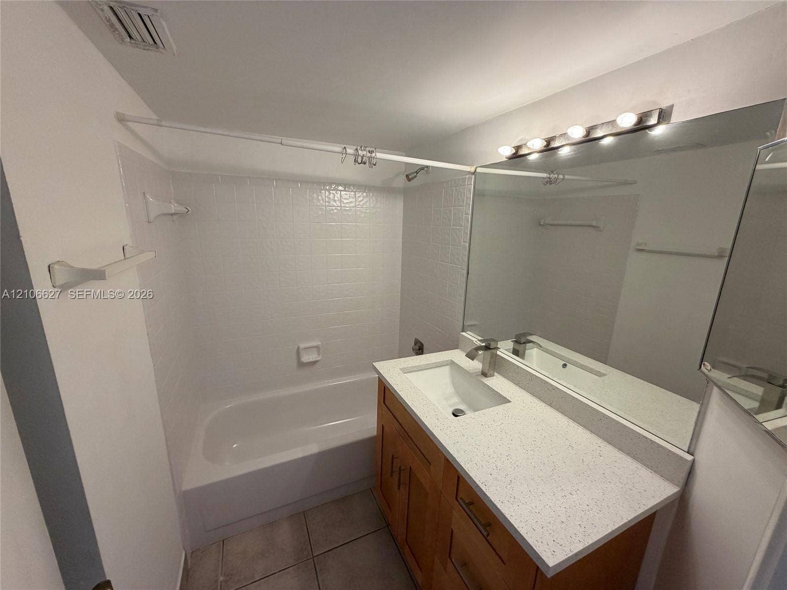 Bathroom featuring wood-finish vanity cabinetry, a stone-flecked composite countertop with an integrated basin, a large wall-mounted vanity mirror, a multi-bulb light fixture, and a tiled shower-tub combination