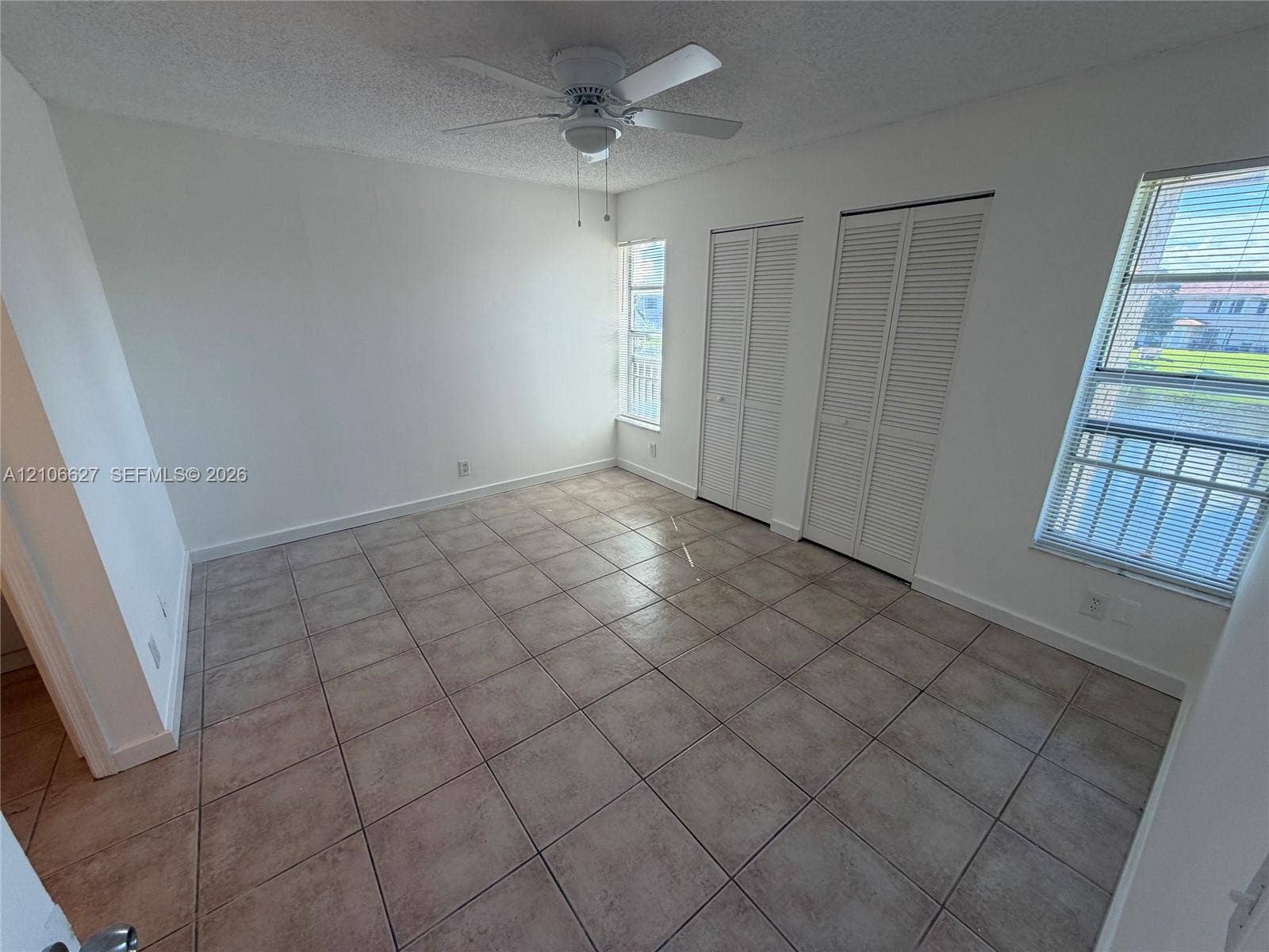 Room featuring neutral tile flooring, dual louvered-door closets, multiple windows, and a ceiling fan