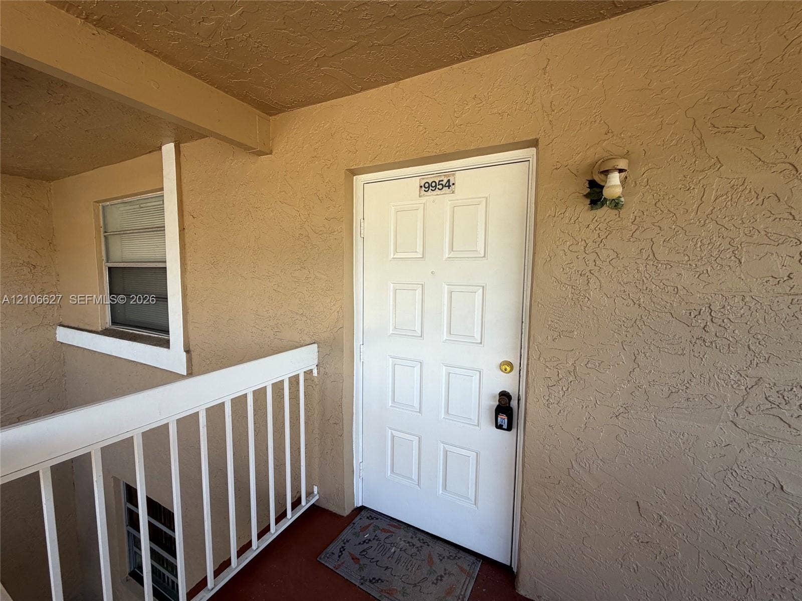 Exterior entryway featuring a white paneled door, textured stucco wall finish, metal railing, and wall-mounted exterior lighting