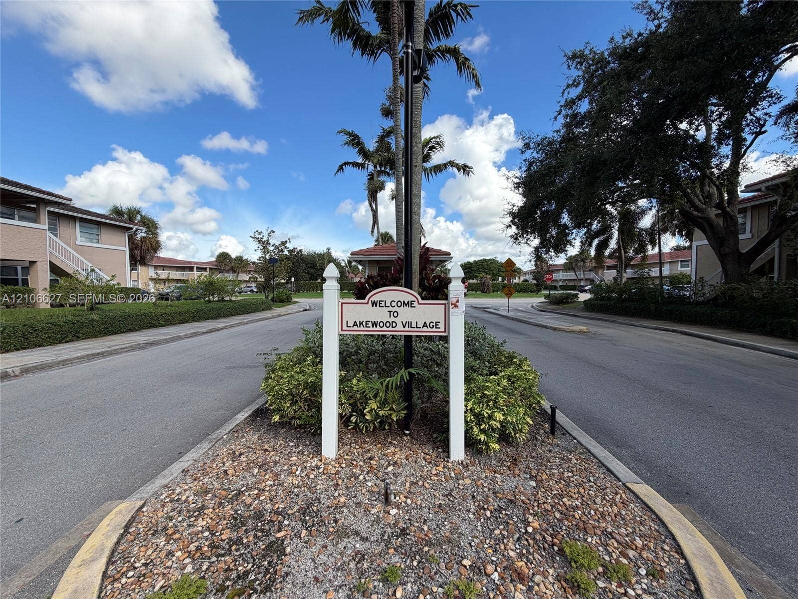 9954 Twin Lakes Drive, #7-O, Coral Springs, Coral Springs, FL 33071 Lakewood Village community signage situated within a landscaped traffic island featuring stone mulch, manicured shrubbery, and mature palm trees