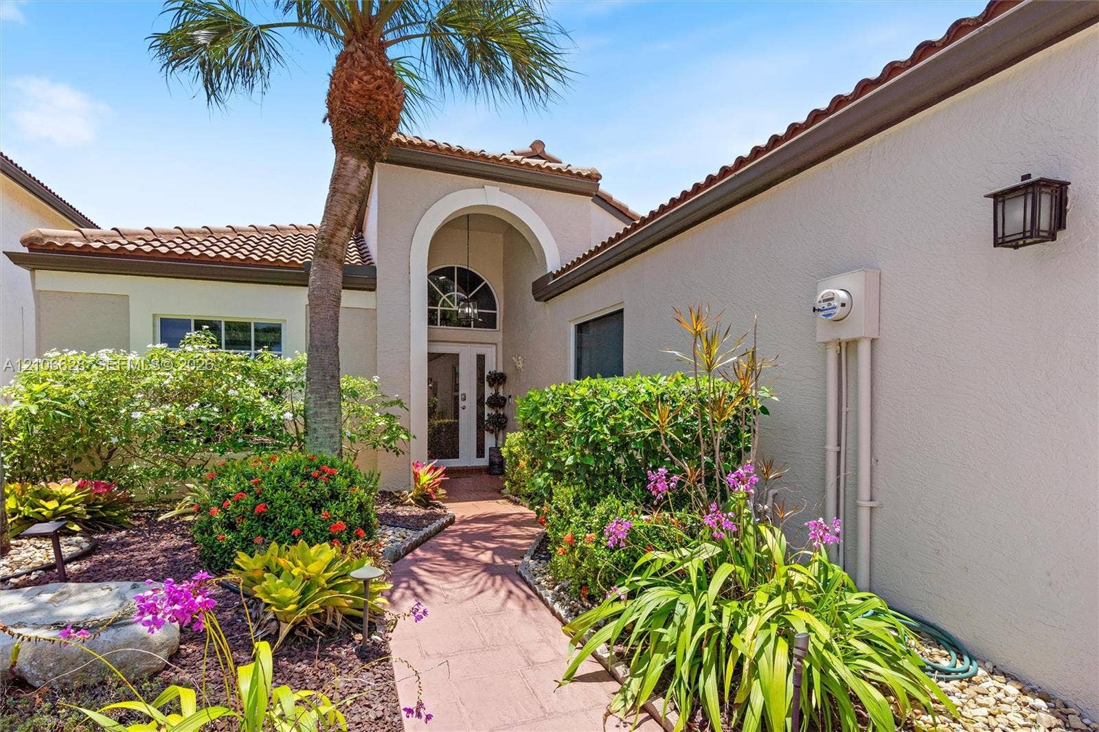 7259 Via Palomar, Boca Raton, FL 33433 Stucco exterior featuring a arched entryway, tile roof, and a paved walkway surrounded by mature landscaping