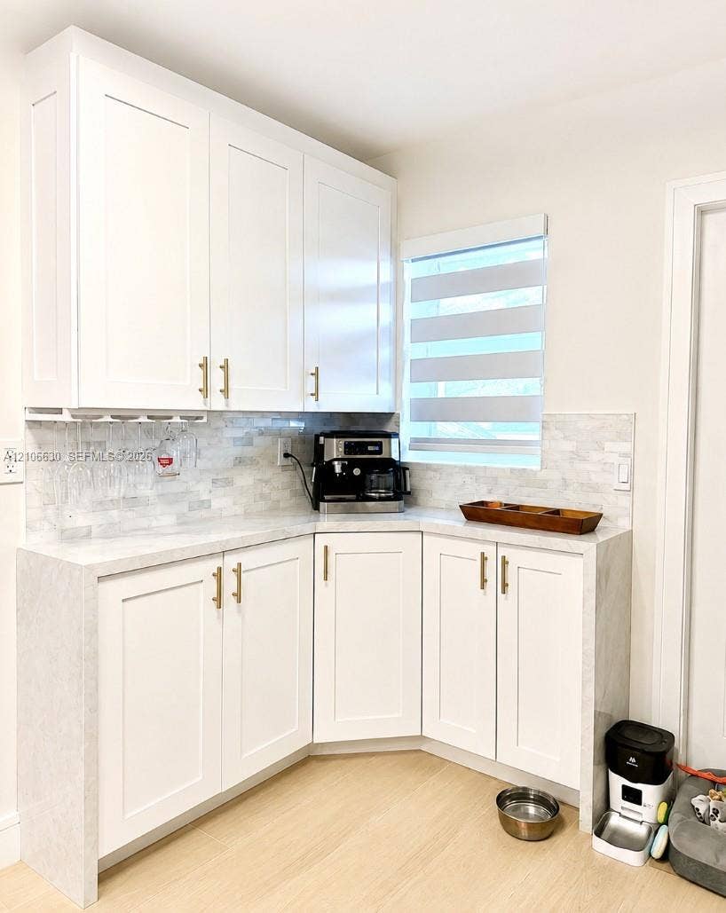 White Shaker-style cabinetry featuring gold-tone hardware, stone-surface countertops, stone-mosaic backsplash, and wood-finish flooring