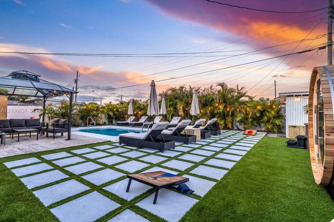 Outdoor living space featuring an in-ground swimming pool, a metal-framed hardtop gazebo, a barrel-style sauna, and a geometric concrete paver patio set in synthetic turf