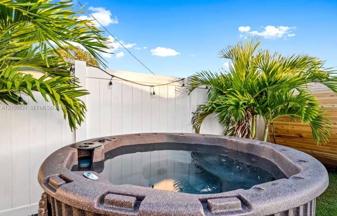Outdoor hot tub featuring a textured exterior finish and built-in cup holders, enclosed by a white vinyl privacy fence and native palm trees
