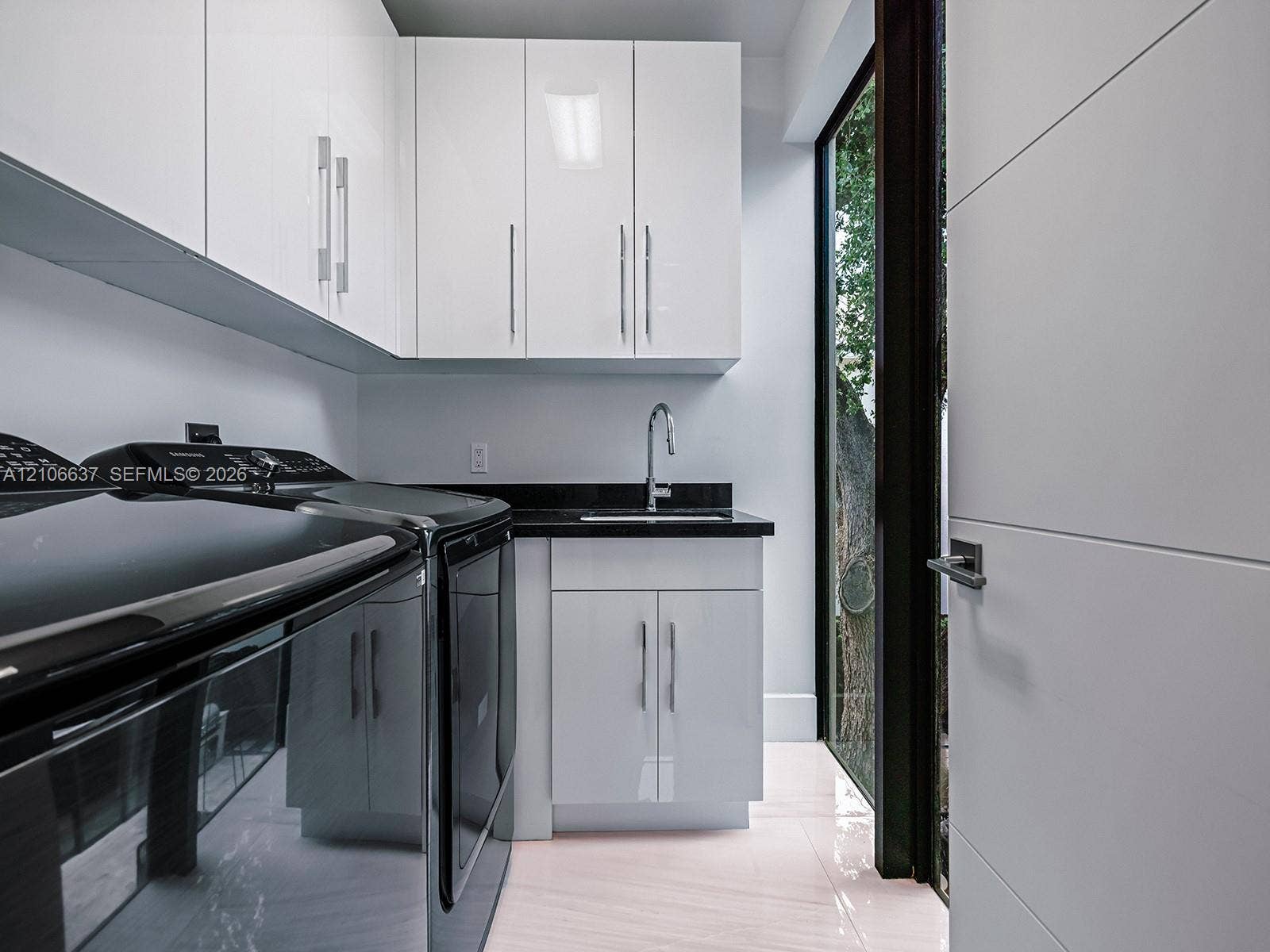 Glossy white cabinetry with vertical pulls, solid-surface dark countertop, undermount sink with gooseneck faucet, light-toned polished flooring, and full-height glass paneled door