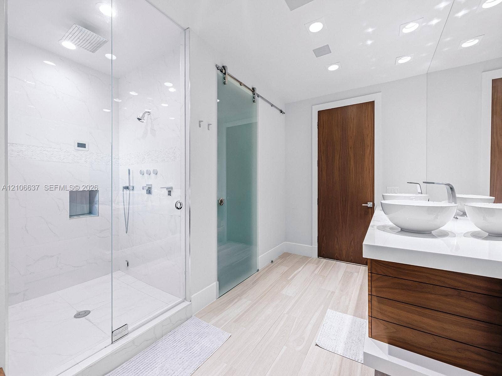 Walk-in glass-enclosed shower with wall-mounted rainfall fixture and recessed niche, dual vessel sinks atop a wood-finish vanity, polished white solid-surface countertop, and wood-finish door