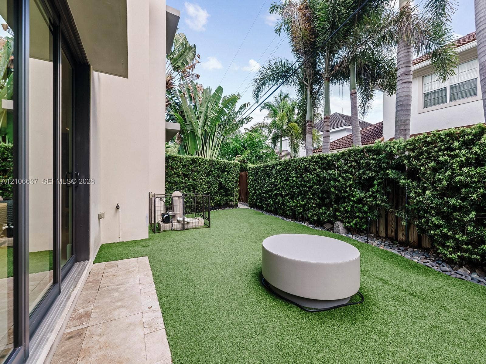 Exterior space featuring an artificial turf lawn, stone-finish patio pavers, perimeter hedges, and sliding glass doors