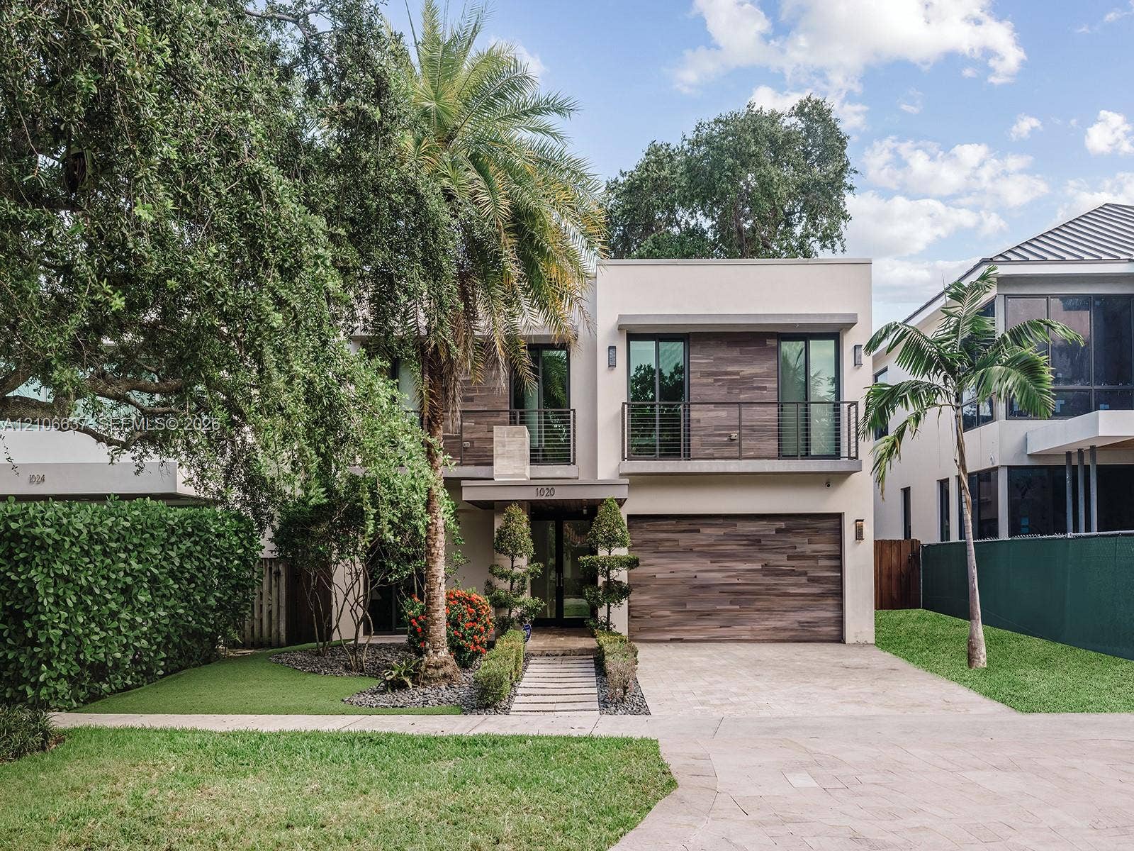 Contemporary two-story exterior featuring smooth stucco siding with wood-tone accents, a stone-paved driveway, a second-story balcony with metal railings, and a horizontal wood-finish garage door