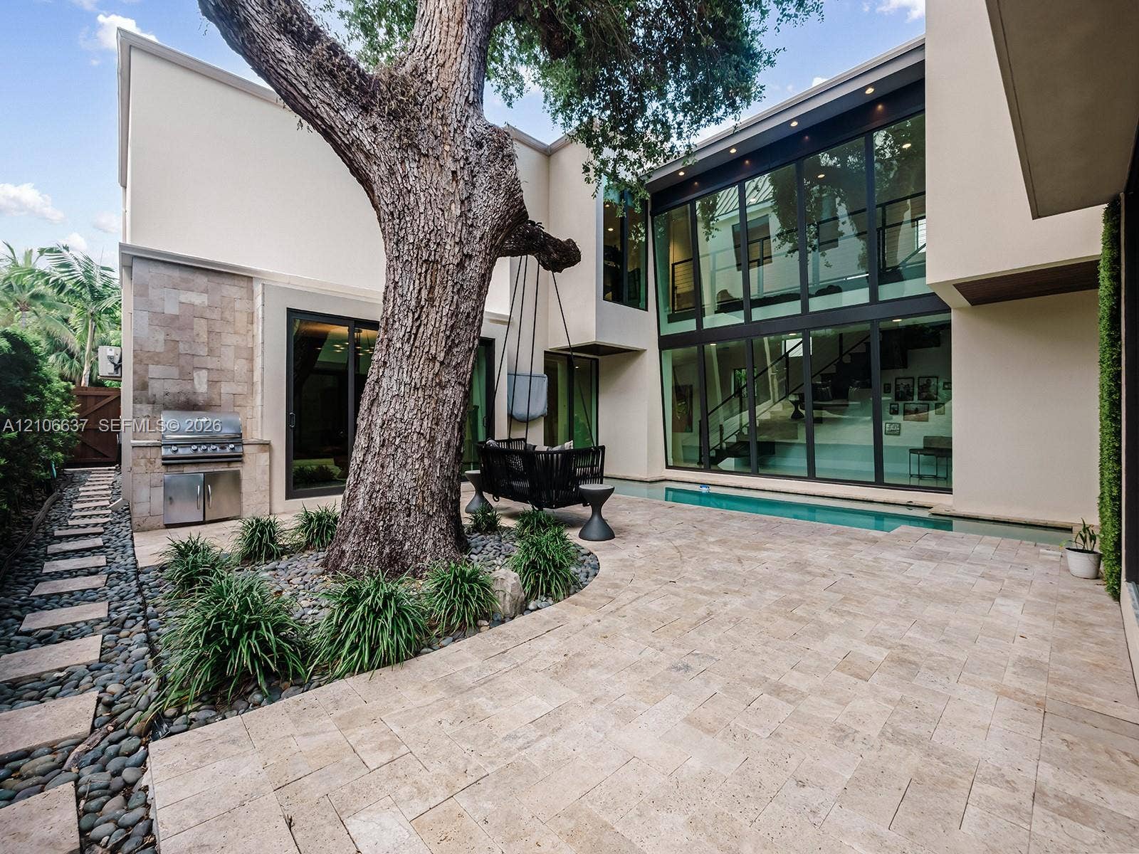 Modern courtyard featuring stone paver flooring, floor-to-ceiling glass windows, a built-in outdoor grill, a mature tree, and a linear swimming pool