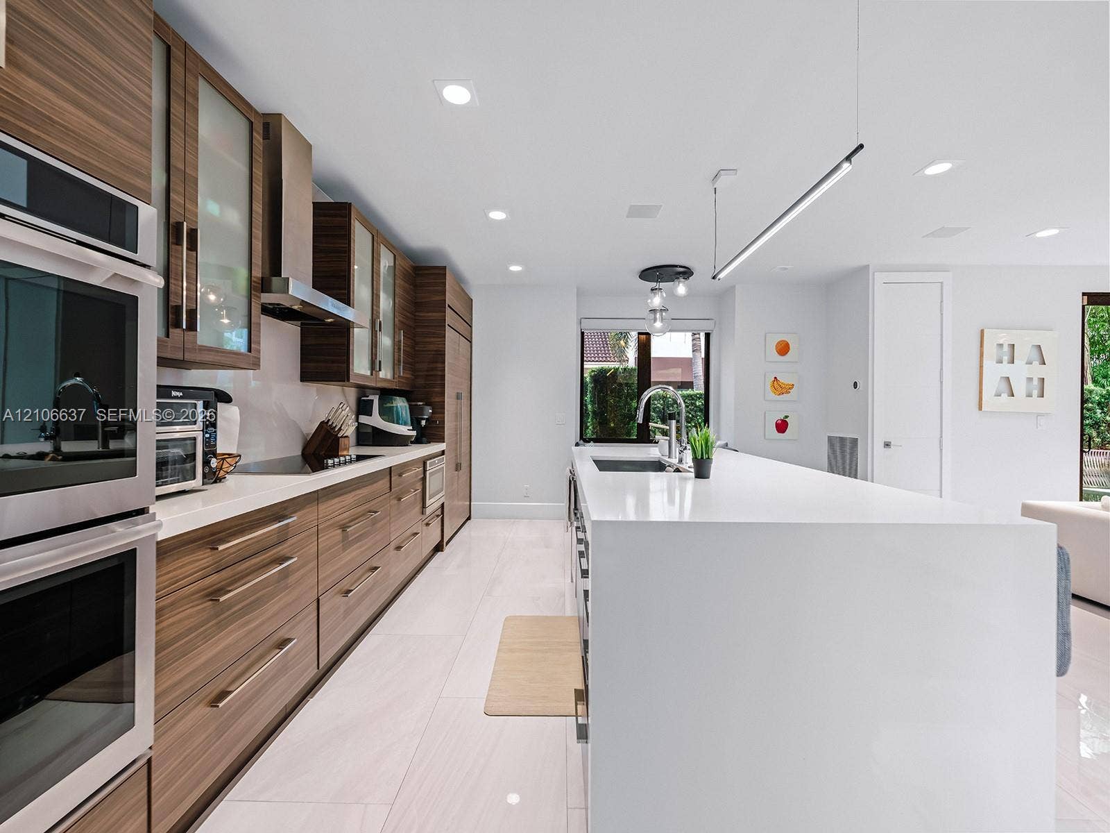 Contemporary kitchen featuring horizontal-grain wood cabinetry, expansive solid-surface island, stainless steel built-in double ovens, integrated stainless steel range hood, and large-format porcelain tile flooring