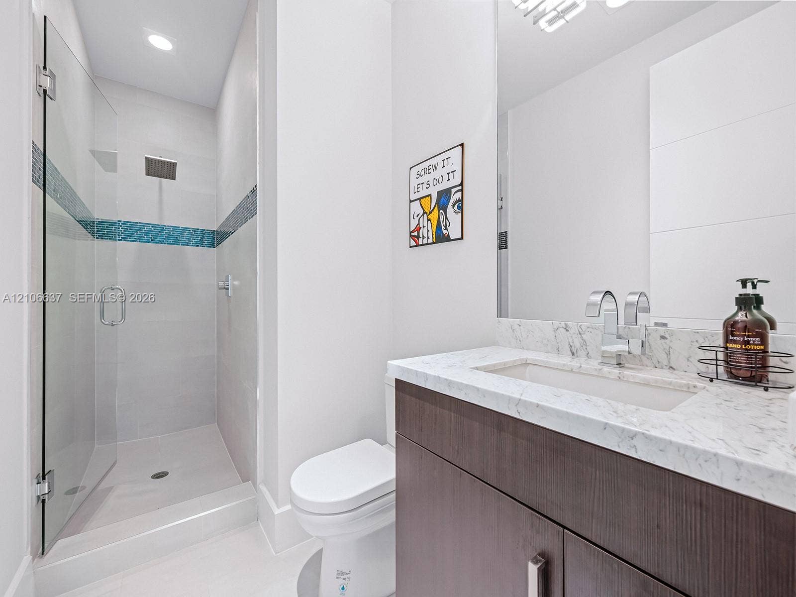Modern bathroom featuring a dark wood-finish vanity, light-toned stone-finish countertop with an integrated rectangular sink, glass-enclosed shower with decorative tile inlay, and chrome fixtures