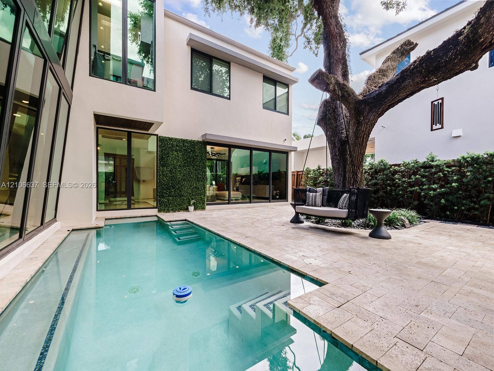 Exterior courtyard featuring an in-ground swimming pool with integrated steps, stone-tile decking, full-height floor-to-ceiling windows, and mature trees