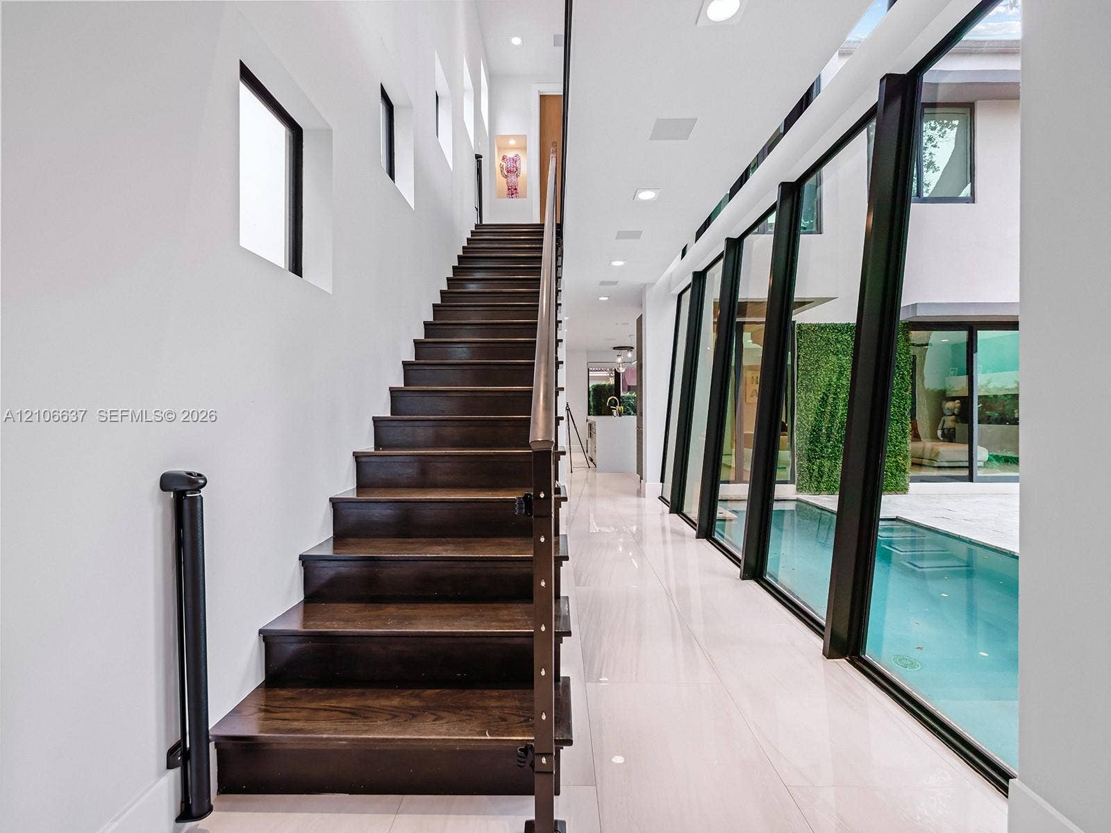 Staircase with dark wood-finish treads and metal railings, large-format tile flooring, recessed ceiling lighting, and floor-to-ceiling black-framed windows overlooking a pool area
