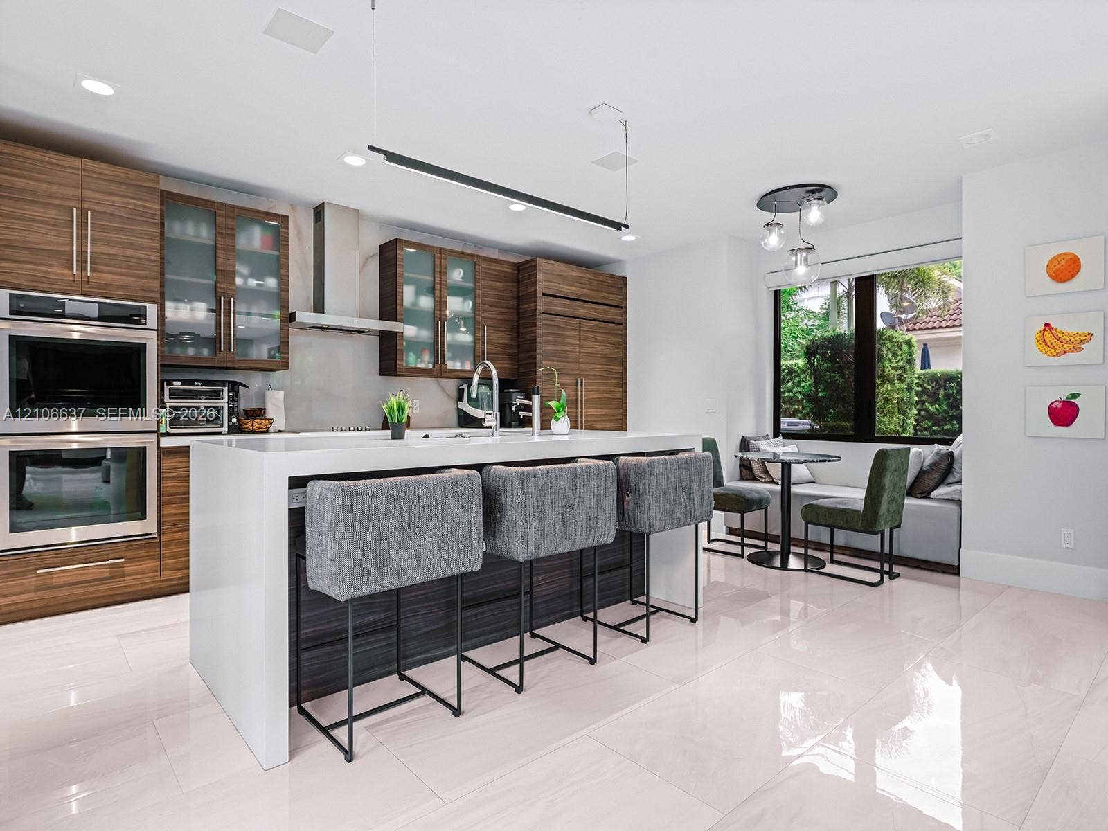 Modern kitchen featuring horizontal-grain wood cabinetry, a central island with a waterfall-edge countertop, built-in double wall ovens, recessed lighting, and large-format tile flooring