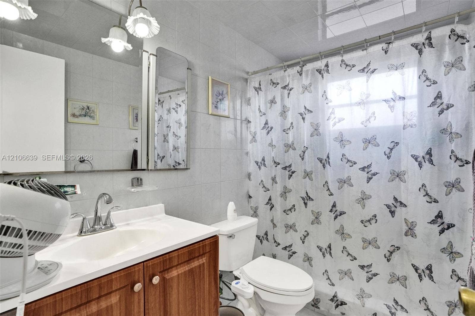 Full-wall vanity mirror with integrated light fixture, wood-finish cabinet, and white countertop featuring a high-arc faucet, complemented by wall-to-wall tiling and a built-in shower and tub combination