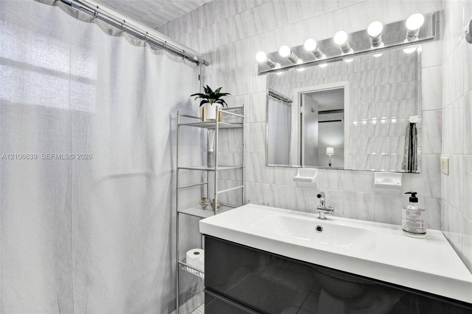 Wall-mounted vanity featuring a glossy dark cabinet, white integrated sink, chrome faucet, full-width mirror with six-bulb light fixture, and floor-to-ceiling wall tile