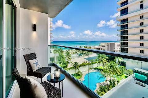 Covered balcony featuring a glass railing, wall-mounted exterior lighting, and overlooking a swimming pool and coastal waters