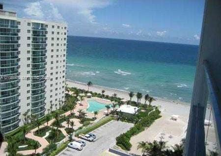 High-rise building with glass-paneled balconies overlooking a coastal landscape featuring a swimming pool, paved walking paths, landscaped grounds, and direct oceanfront access