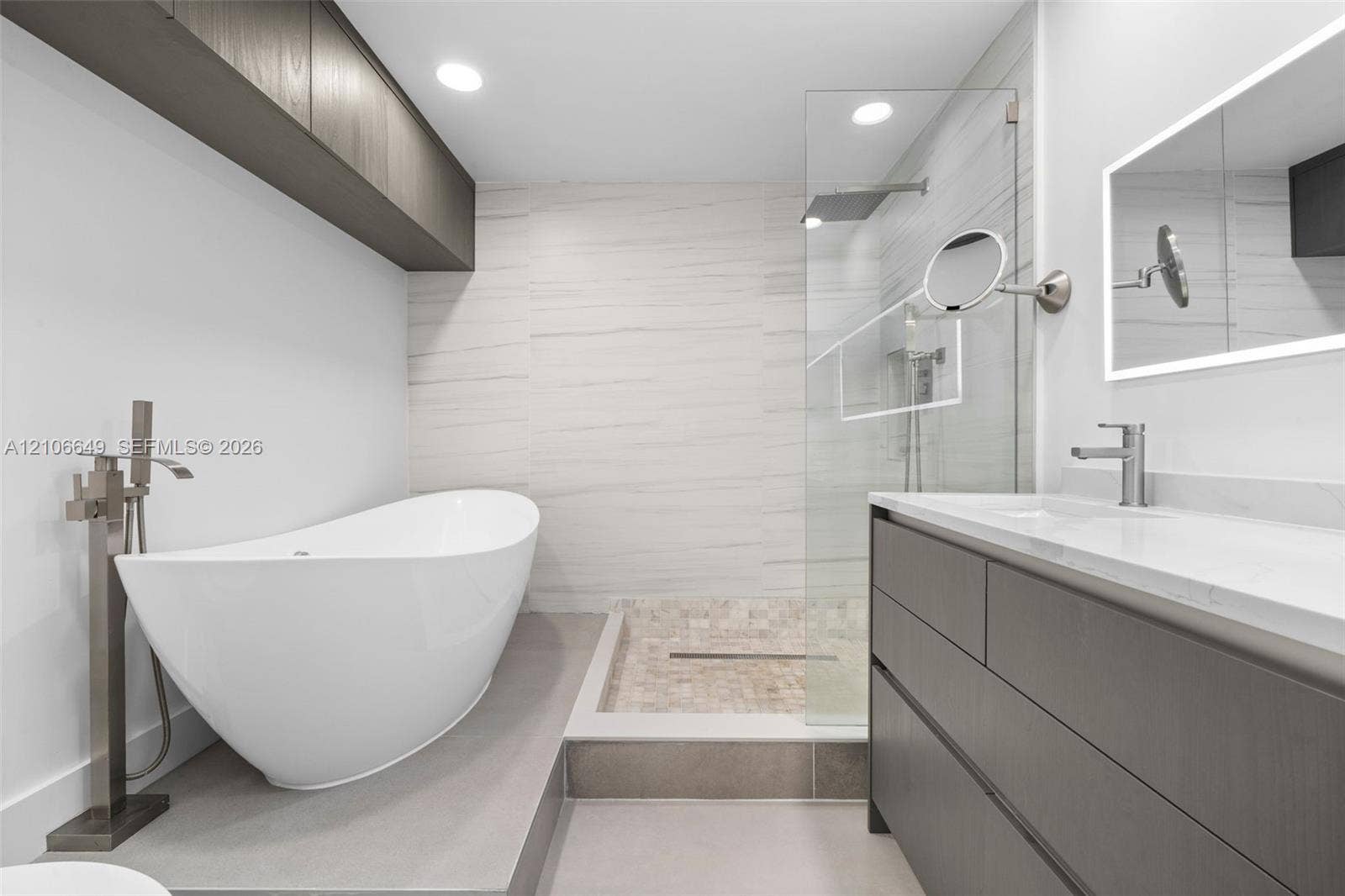 216 Poinciana Drive, #206, Sunny Isles Beach, FL 33160 Freestanding soaking tub with floor-mounted faucet, walk-in glass-enclosed shower featuring mosaic stone flooring, dark wood-finish cabinetry, illuminated wall-mounted mirror, and recessed lighting