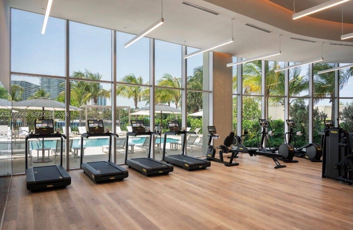 Fitness center featuring floor-to-ceiling windows, wood-finish flooring, linear suspended LED lighting, and recessed ceiling architecture
