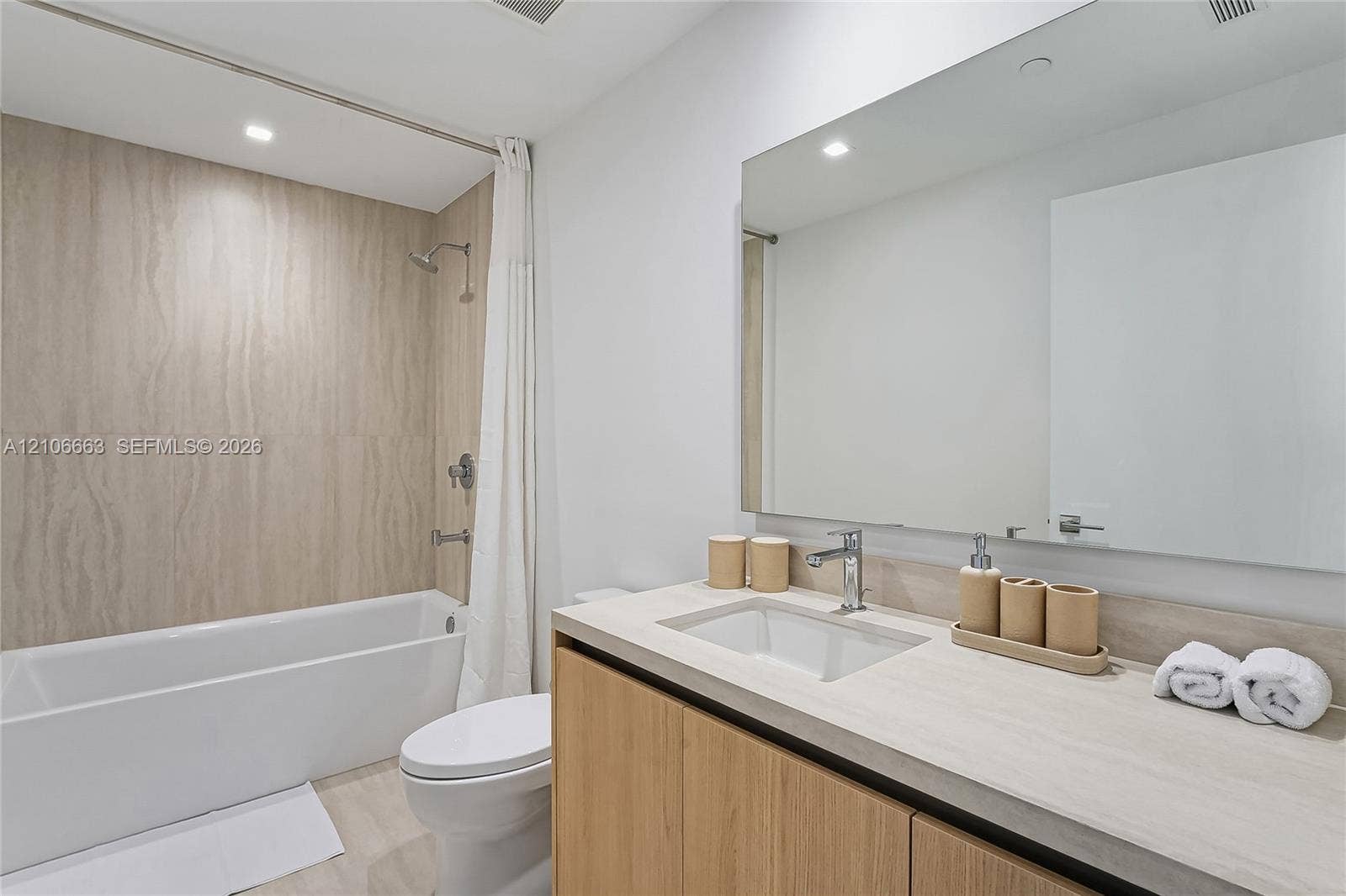 Bathroom featuring a vanity with light-tone wood-finish cabinetry, a solid-surface countertop with an integrated sink, a large frameless mirror, and a soaking tub with neutral stone-finish tile surround