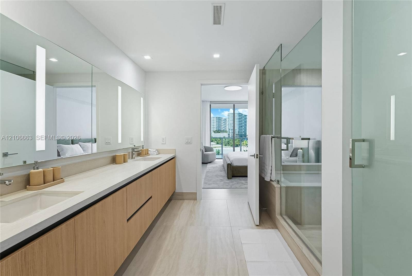 Dual-sink vanity with light wood-finish cabinetry, solid-surface countertop, full-width vanity mirror with integrated vertical lighting, and glass-enclosed shower with tile flooring