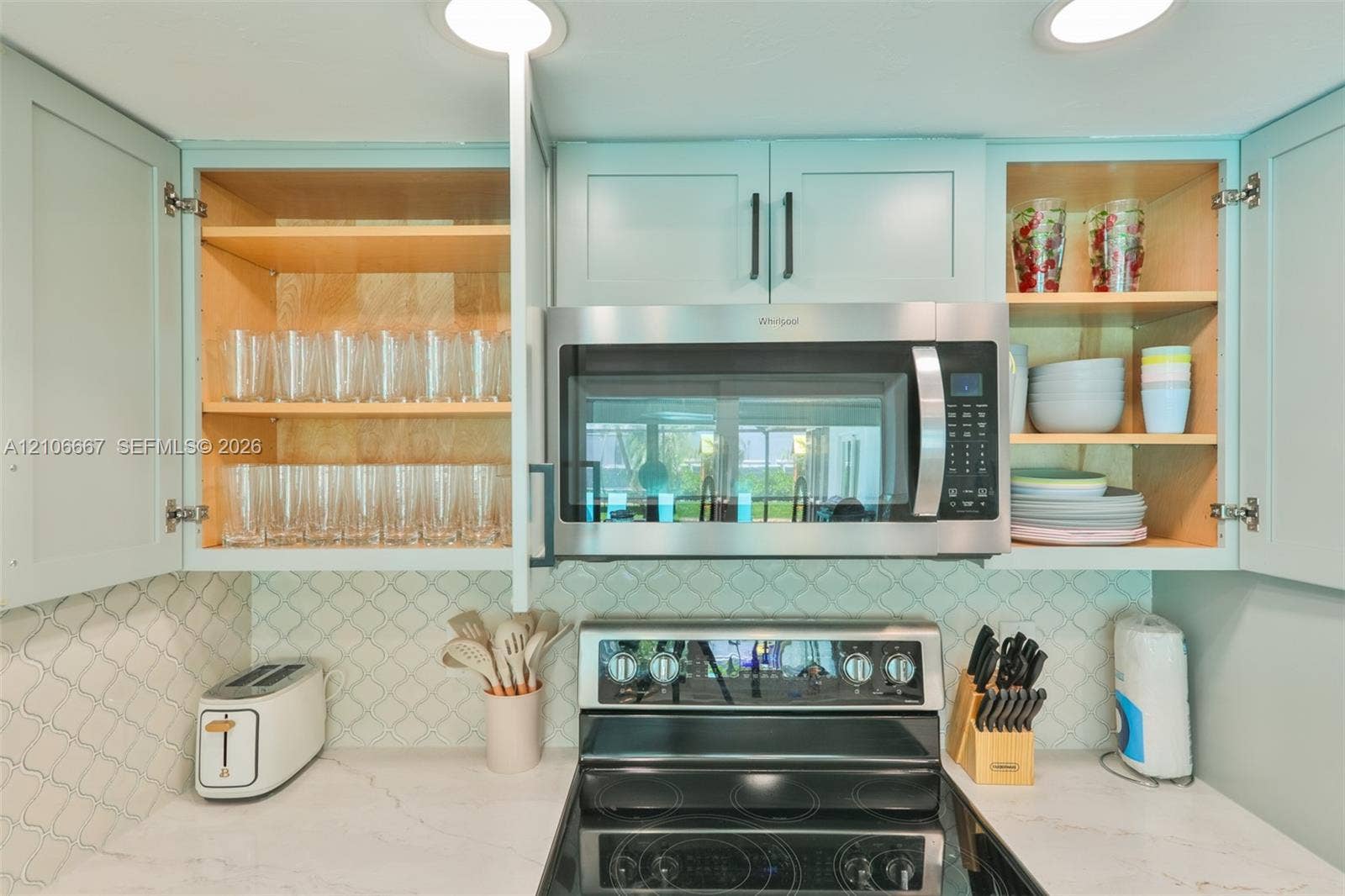 209 Bayshore Drive, Cape Coral, FL 33904 Custom cabinetry with light-toned finish, stainless steel over-the-range microwave, smooth-top electric range, patterned tile backsplash, and light-toned solid surface countertops with integrated recessed lighting