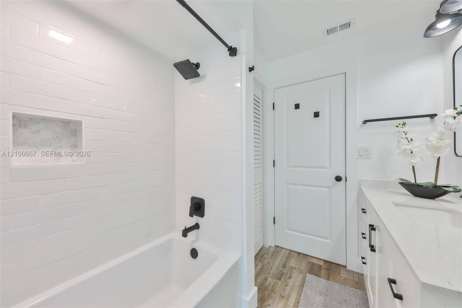 209 Bayshore Drive, Cape Coral, FL 33904 Bathroom featuring a white subway-tiled tub enclosure with an integrated recessed niche, matte black fixtures, white vanity with stone-finish countertop, and wood-finish flooring