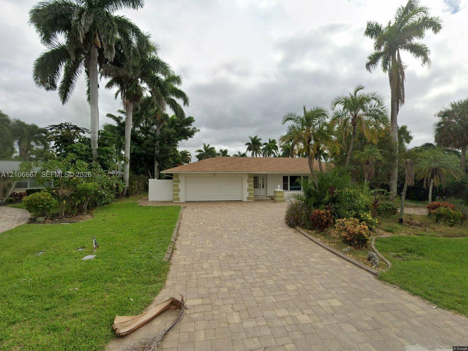 209 Bayshore Drive, Cape Coral, FL 33904 Single-story residence featuring a shingle roof, paved stone driveway, attached two-car garage, and established landscaping with mature palm trees