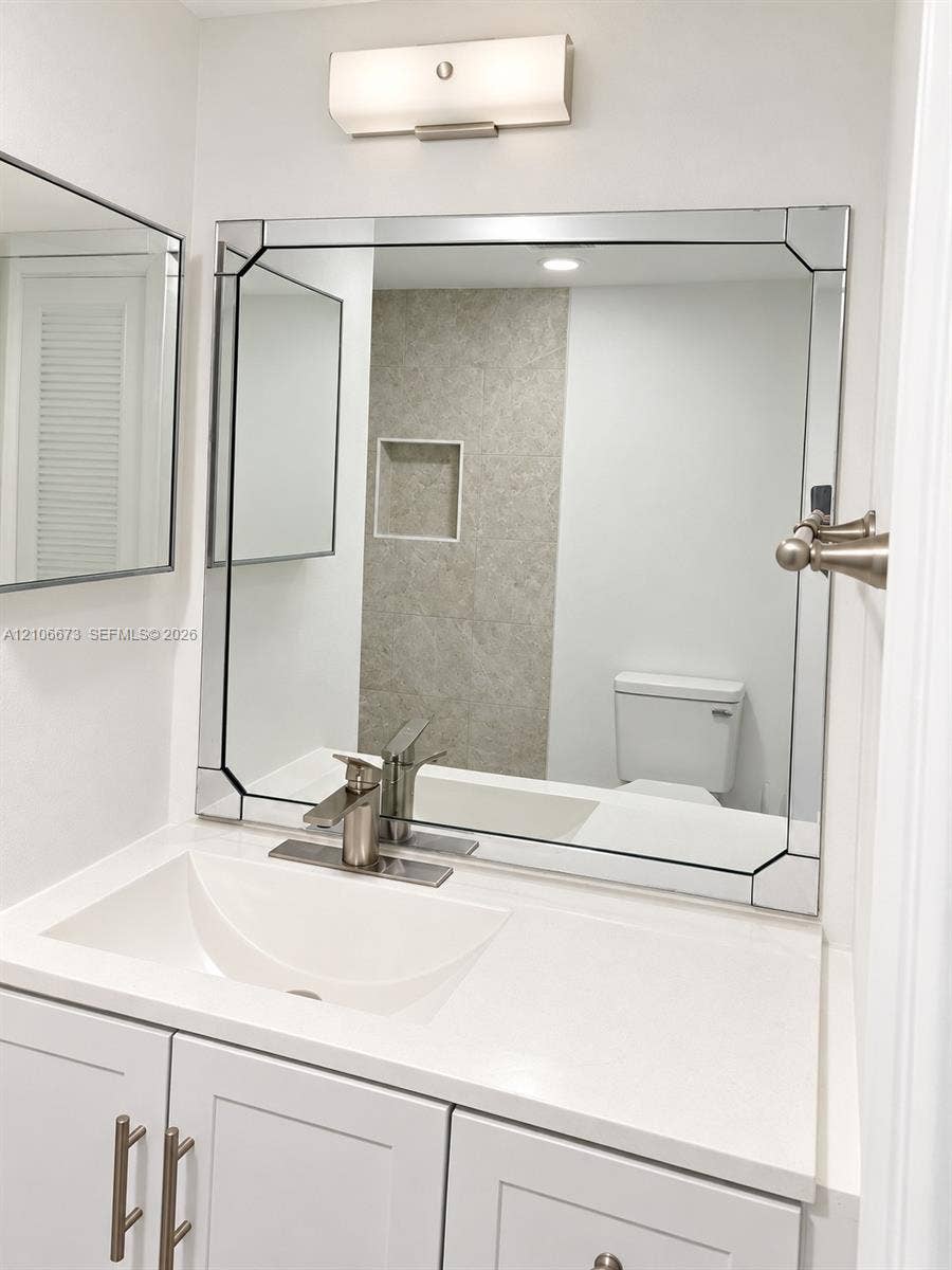 704 SE 2nd Avenue, #440, Deerfield Beach, Deerfield Beach, FL 33441 White vanity with a solid-surface countertop and integrated sink, framed beveled wall mirror, brushed-nickel faucet, satin-finish wall lighting, and tiled wall niche