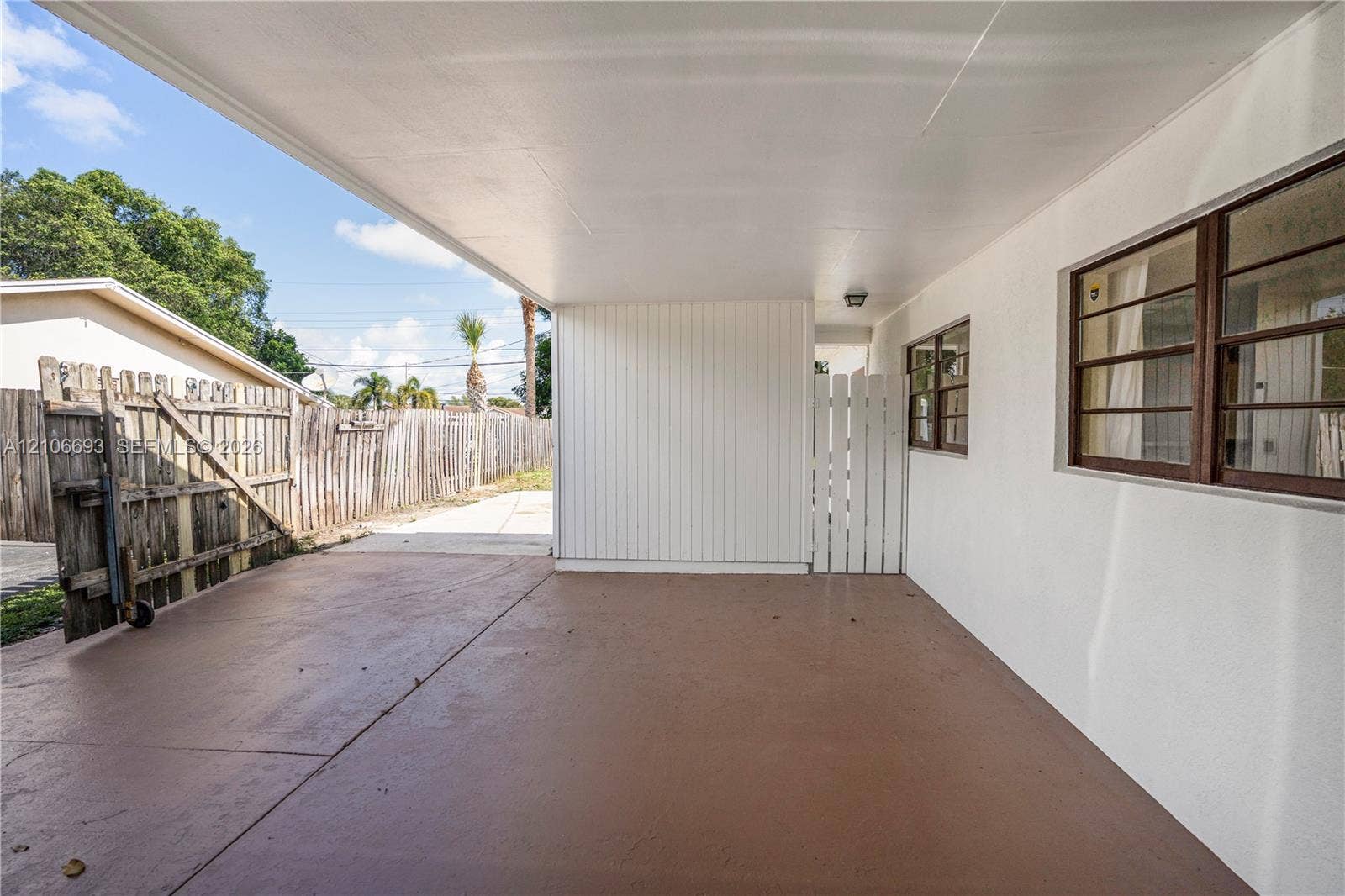 1459 42nd Street, West Palm Beach, FL 33407 Covered concrete carport featuring white vertical siding, single-hung windows with metal frames, and a white privacy gate