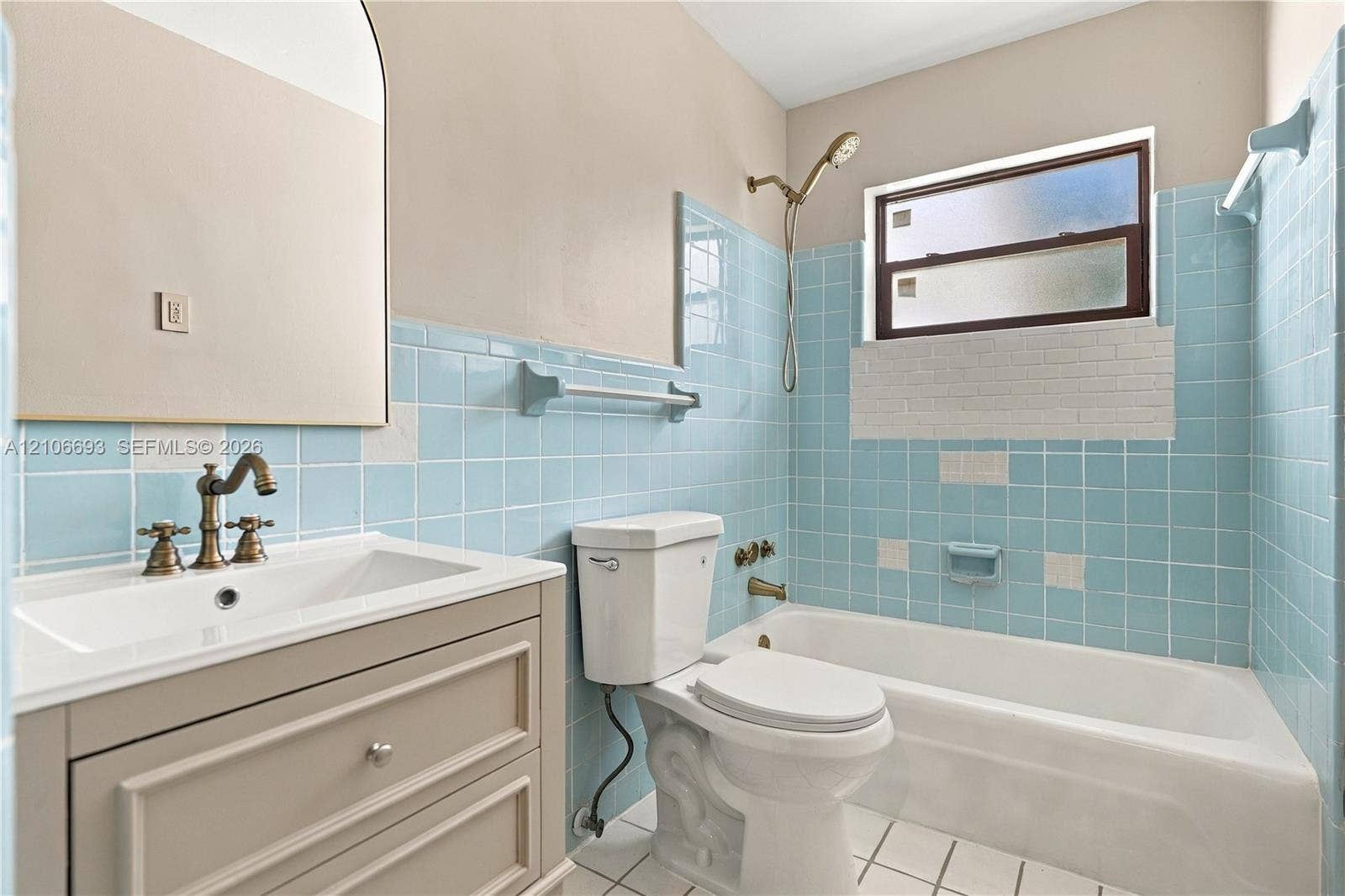 1459 42nd Street, West Palm Beach, FL 33407 Bathroom featuring blue tile wainscoting, vanity with integrated sink, brass-finish faucet and shower fixtures, arched wall mirror, and bathtub with tile surround