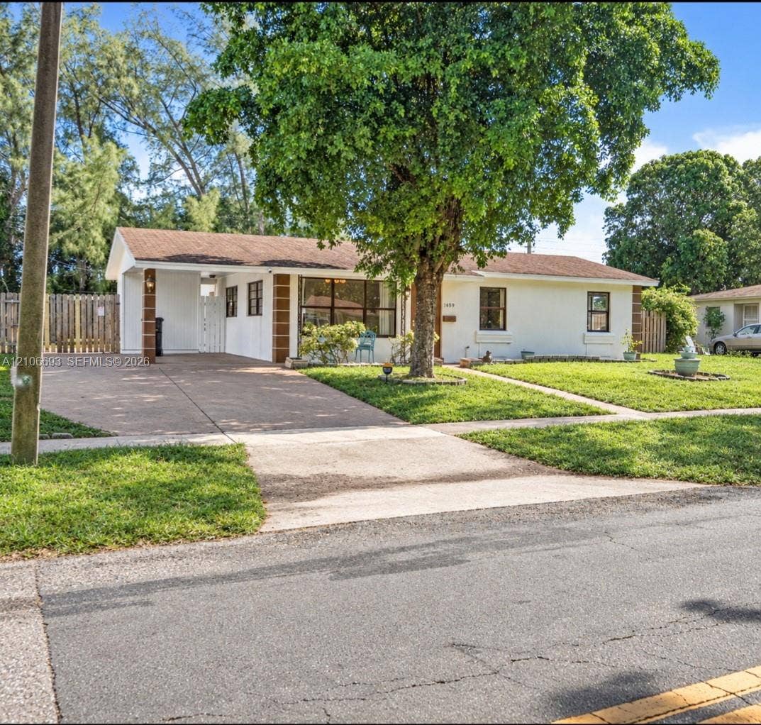 1459 42nd Street, West Palm Beach, FL 33407 Single-story exterior featuring light-toned stucco siding, brown masonry accents, an attached covered carport, a concrete driveway, and mature landscaping