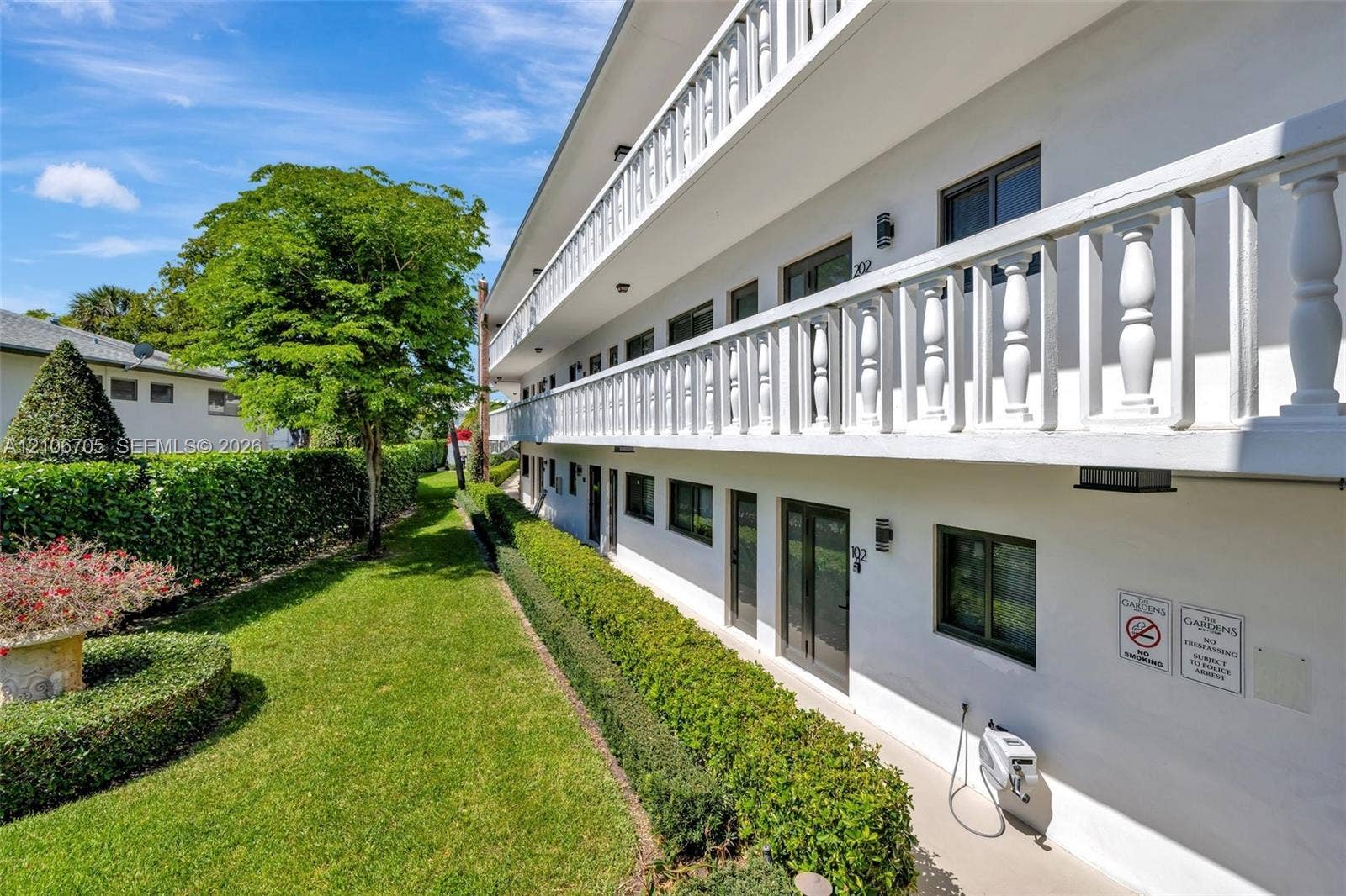 Multi-story exterior featuring white stucco siding, a wrap-around balcony with white balustrades, manicured hedge landscaping, and mature trees
