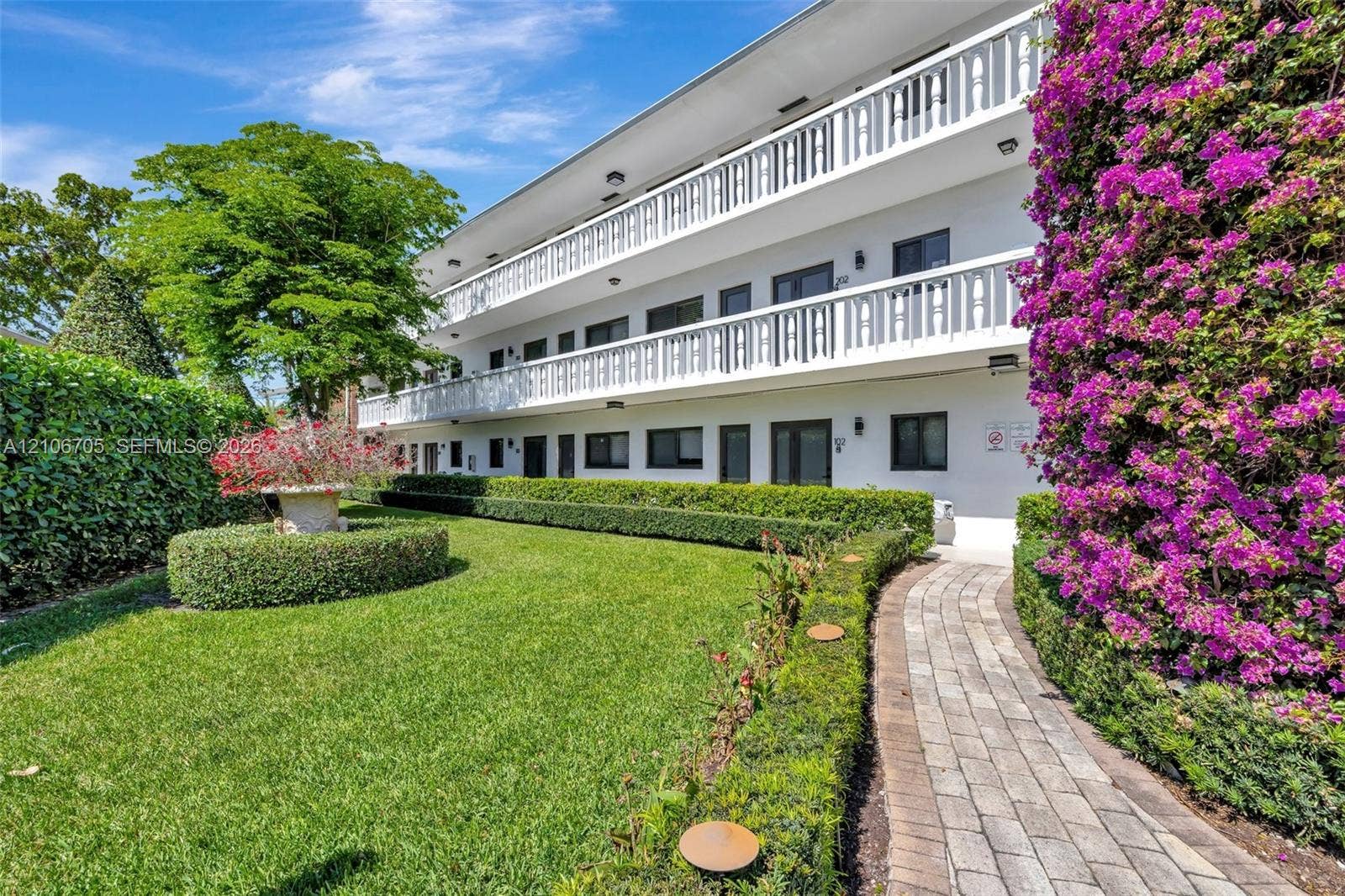 Multi-story building exterior featuring white stucco siding, tiered balconies with decorative railings, manicured hedges, and a paver stone walkway