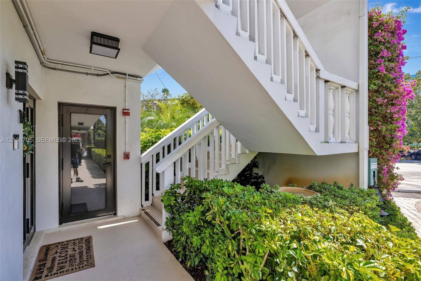 Exterior entryway featuring a glass-panel door, white balustrade staircase, wall-mounted exterior lighting, and mature landscape shrubbery