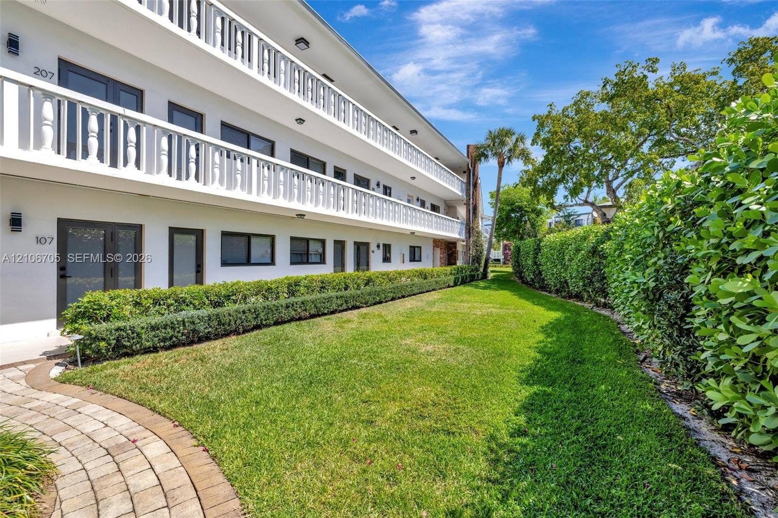 Multi-story building exterior featuring white balustrade railings, paved stone walkways, manicured lawn, and mature privacy hedging