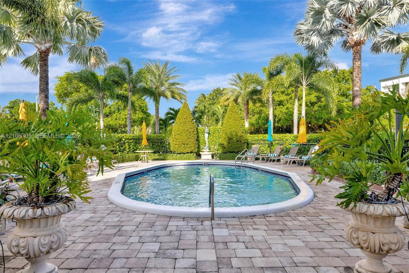 Oval swimming pool with a stone paver deck, surrounding mature palm trees, sculpted hedges, and classical-style decorative urns