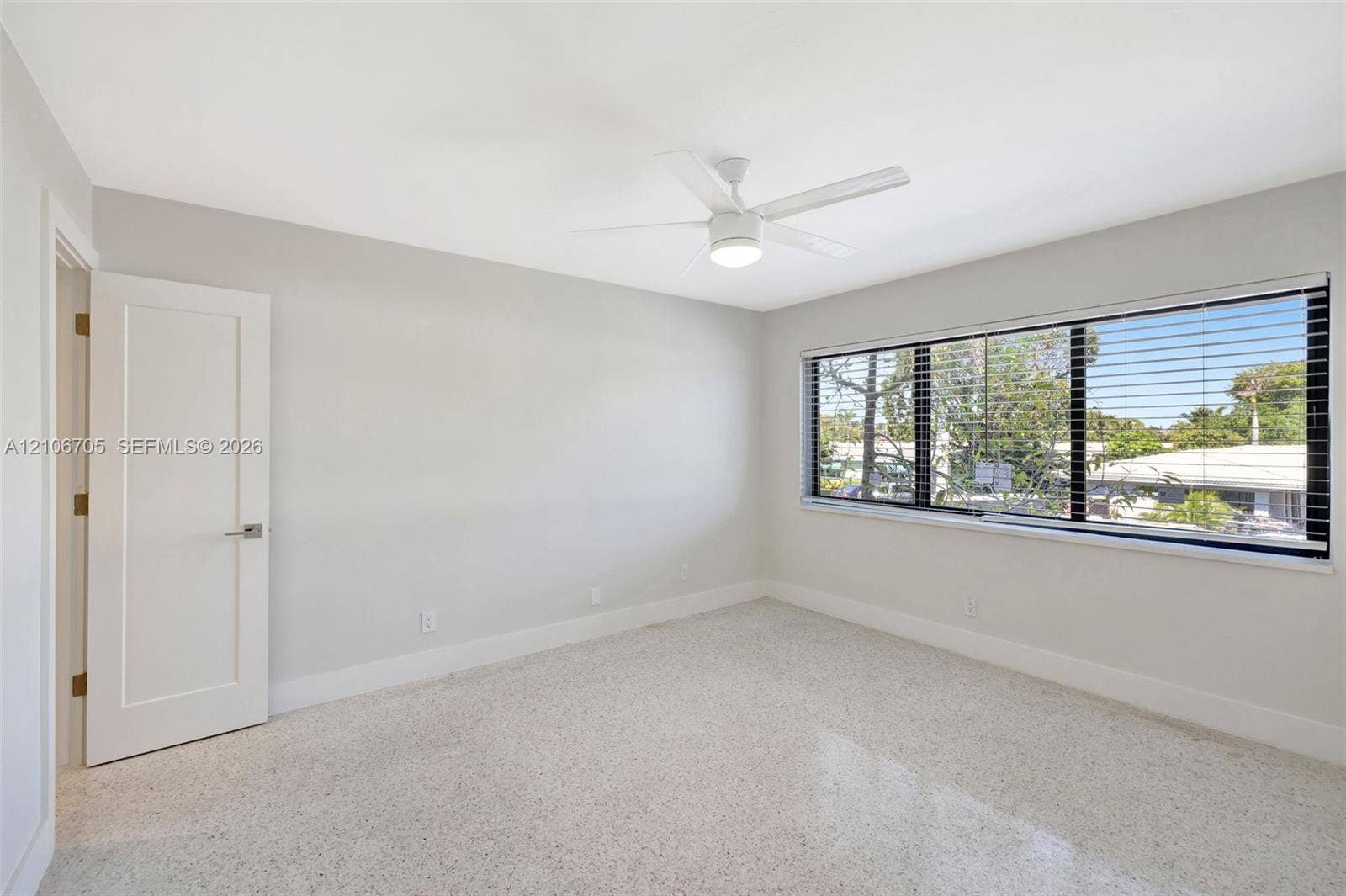 Neutral-toned room featuring a wide window with integrated horizontal blinds, a white ceiling fan with light fixture, and light-colored speckled flooring