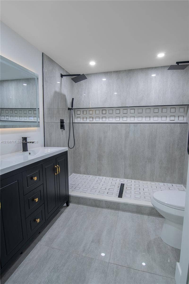 6811 Thomas Street, Hollywood, FL 33024 Bathroom featuring a dark wood-finish vanity with gold-tone hardware, white rectangular sink, grey stone-finish tile flooring, and a walk-in shower with dual rain-style showerheads and decorative tile inlay