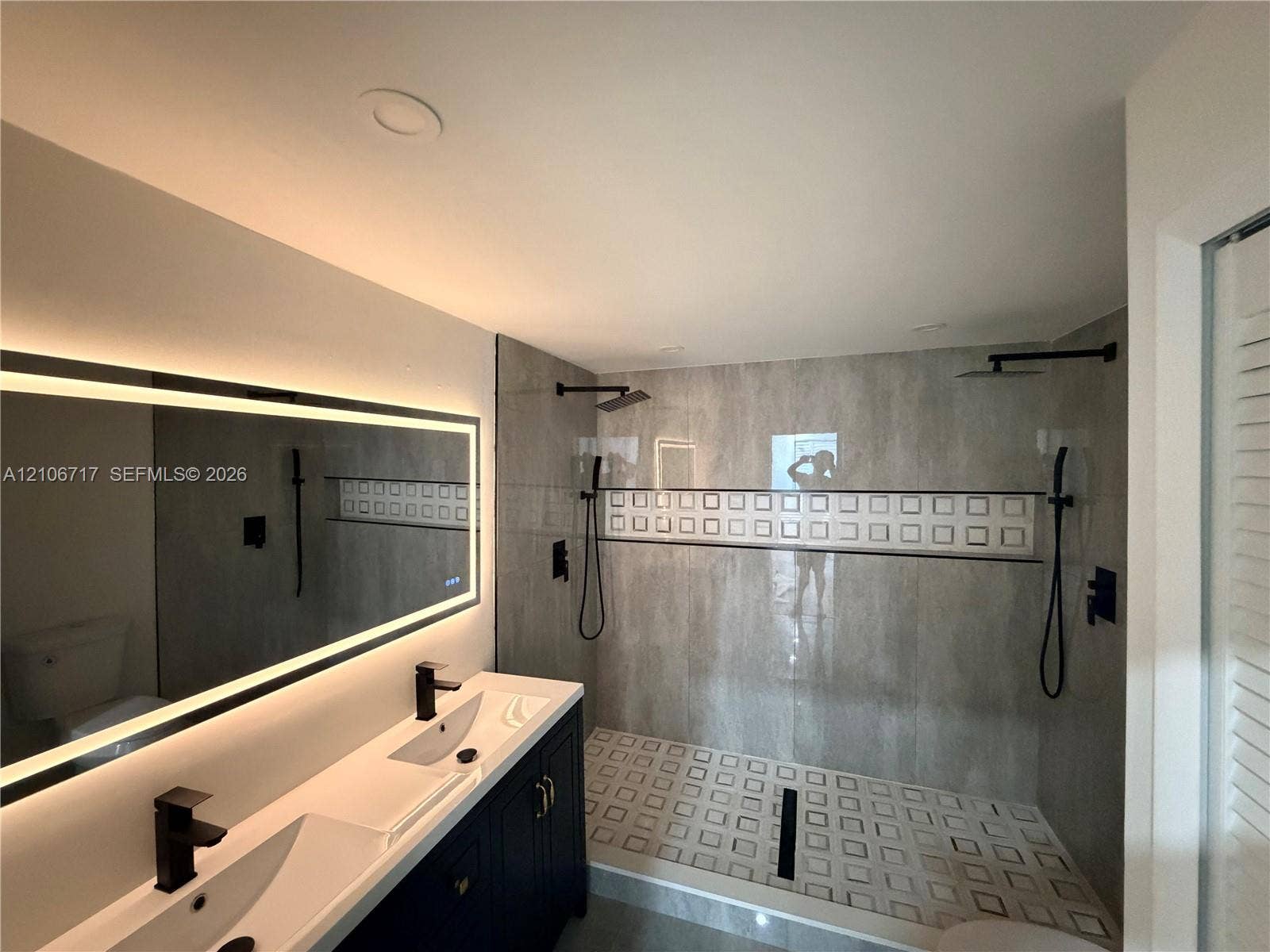 6811 Thomas Street, Hollywood, FL 33024 Double vanity featuring integrated sinks, matte black faucets, and a large backlit wall mirror, paired with a walk-in shower showcasing oversized stone-finish wall tile, a decorative square-patterned inlay, and dual shower heads