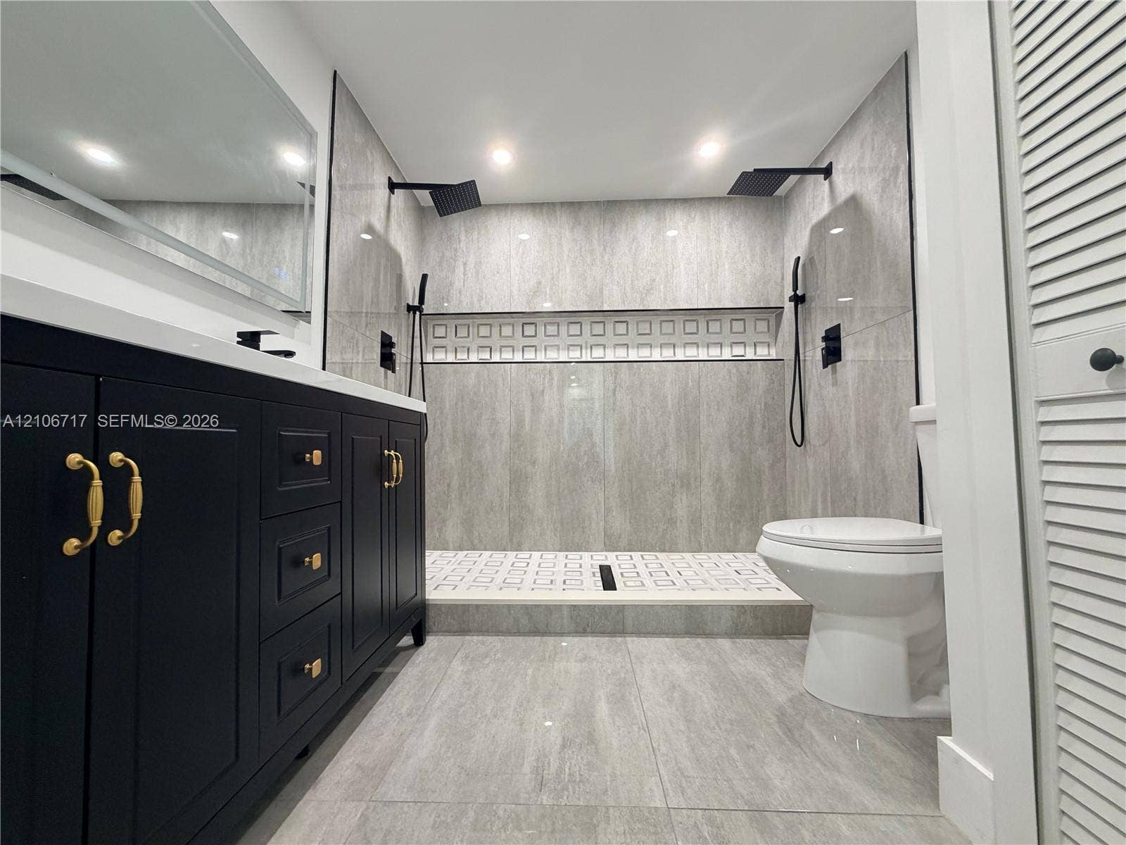 6811 Thomas Street, Hollywood, FL 33024 Dark cabinetry with gold-toned hardware, stone-finish flooring, walk-in shower with dual matte black rainfall showerheads, and decorative tile inlay