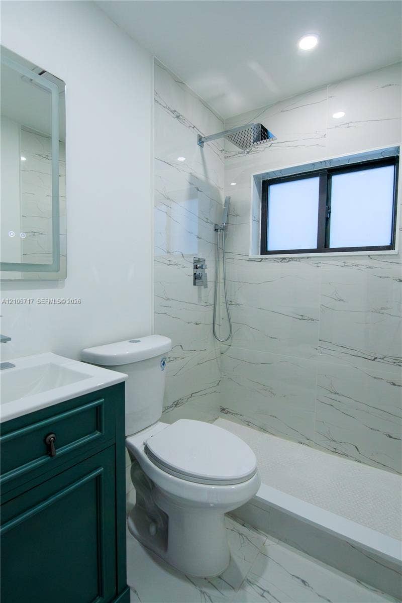 6811 Thomas Street, Hollywood, FL 33024 Bathroom featuring a green-toned vanity with a white countertop, a walk-in shower with large-format veined wall tiles, a rainfall showerhead, and a rectangular LED-backlit mirror