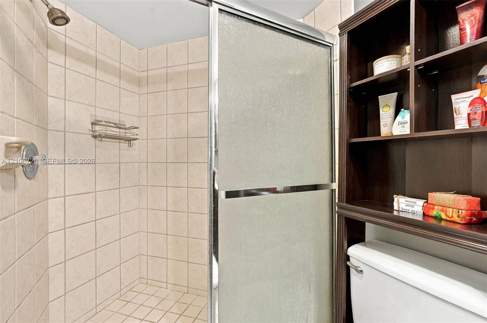 152 Weybridge Circle, #C, Royal Palm Beach, FL 33411 Walk-in shower featuring a sliding glass door, neutral tile surround, wall-mounted caddy, and overhead showerhead, complemented by built-in dark wood shelving above the toilet