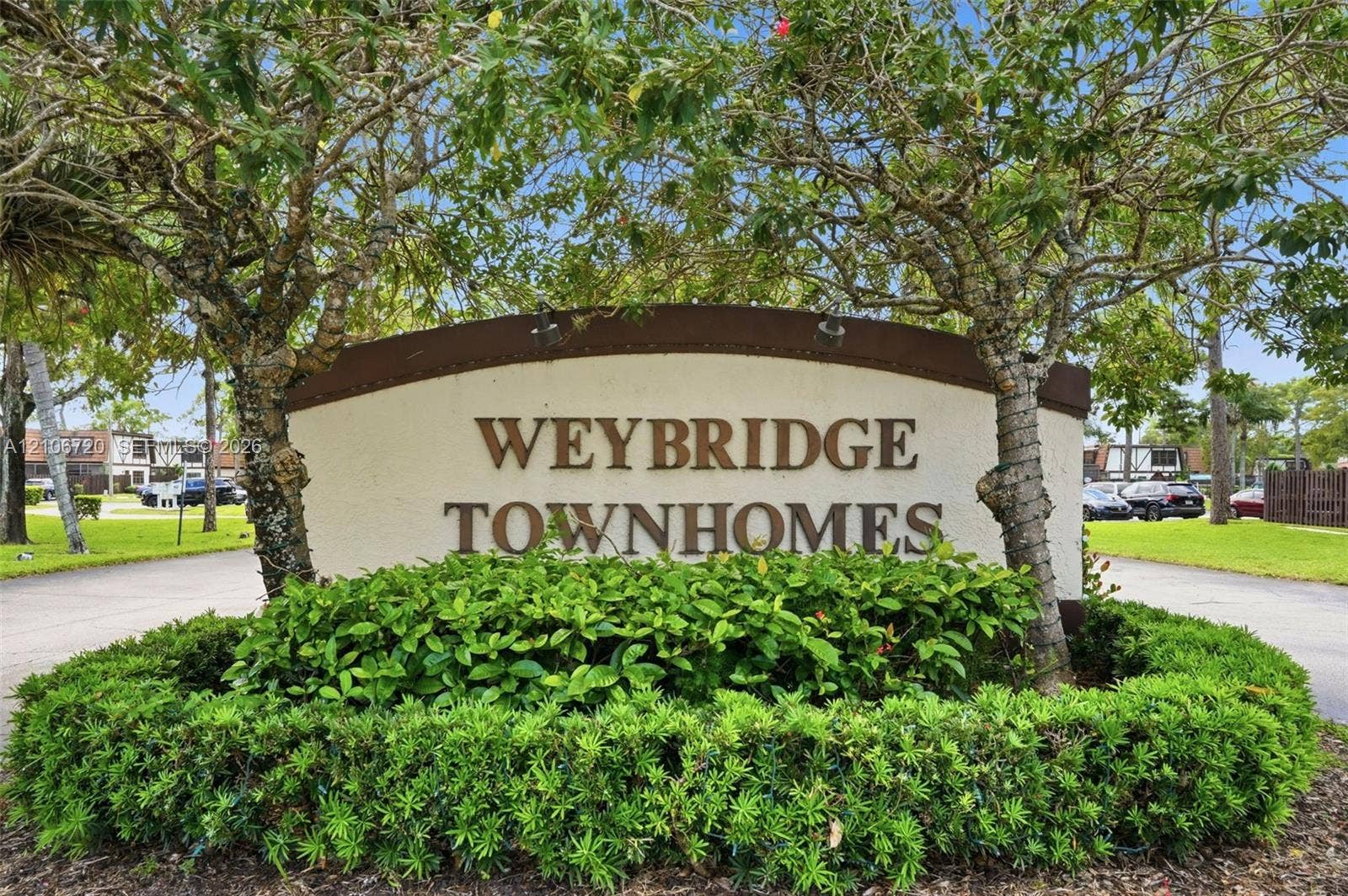 152 Weybridge Circle, #C, Royal Palm Beach, FL 33411 Weybridge Townhomes monument sign featuring dimensional lettering, decorative arch-top trim, and integrated landscape lighting, surrounded by manicured shrubbery and mature canopy trees