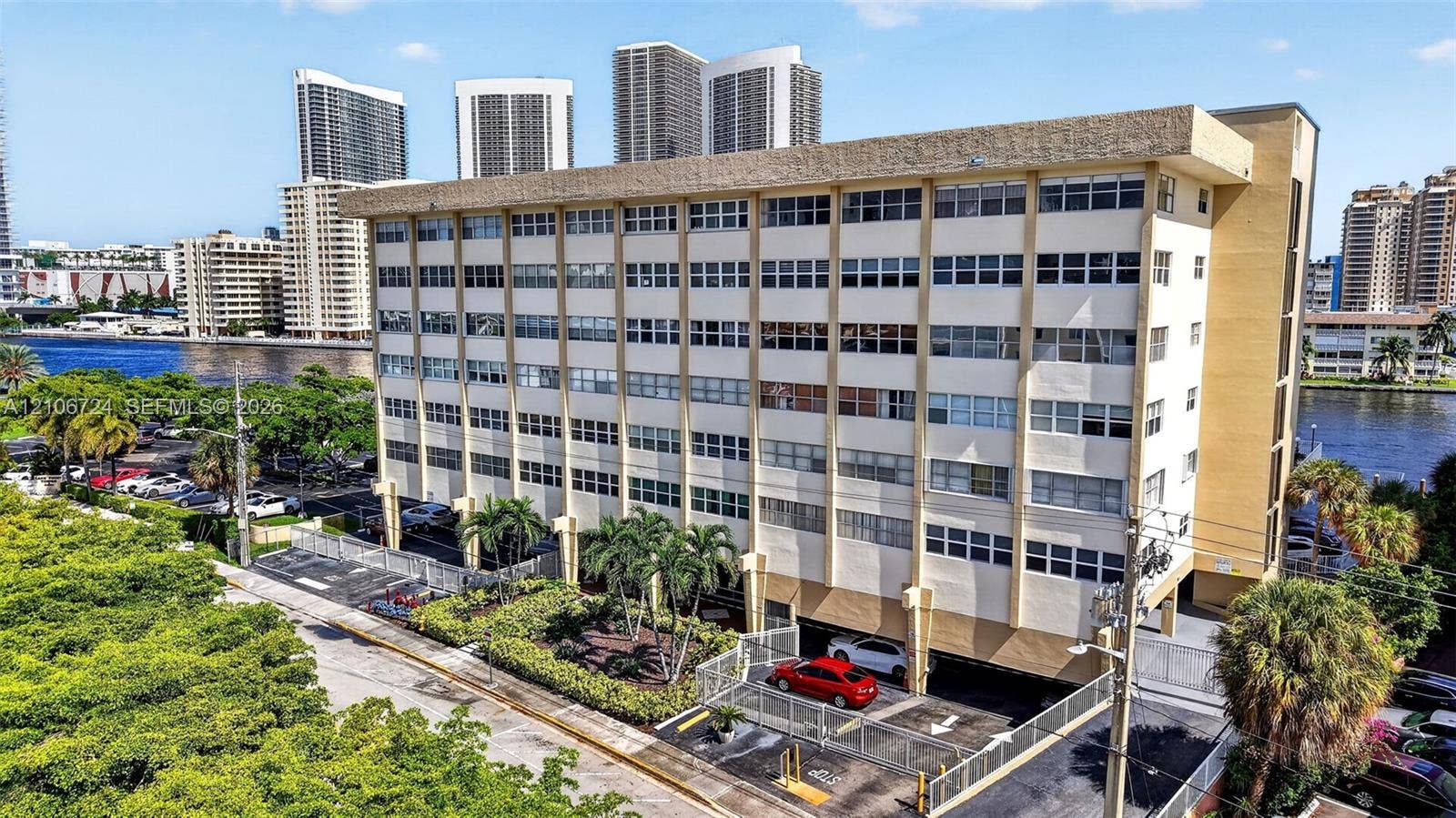 111 Golden Isles Drive, #G12, Hallandale Beach, FL 33009 Multi-story residential building featuring a concrete facade, expansive window rows, ground-level covered parking, and perimeter landscaping