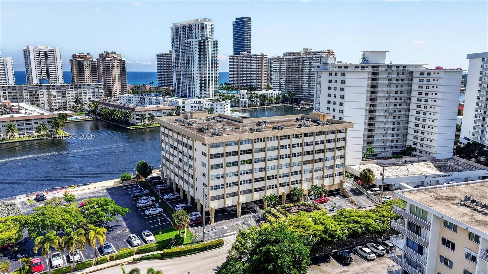 111 Golden Isles Drive, #G12, Hallandale Beach, FL 33009 Aerial perspective of a multi-story residential building featuring covered ground-level parking, landscaped perimeter greenery, and direct water frontage along an inlet canal