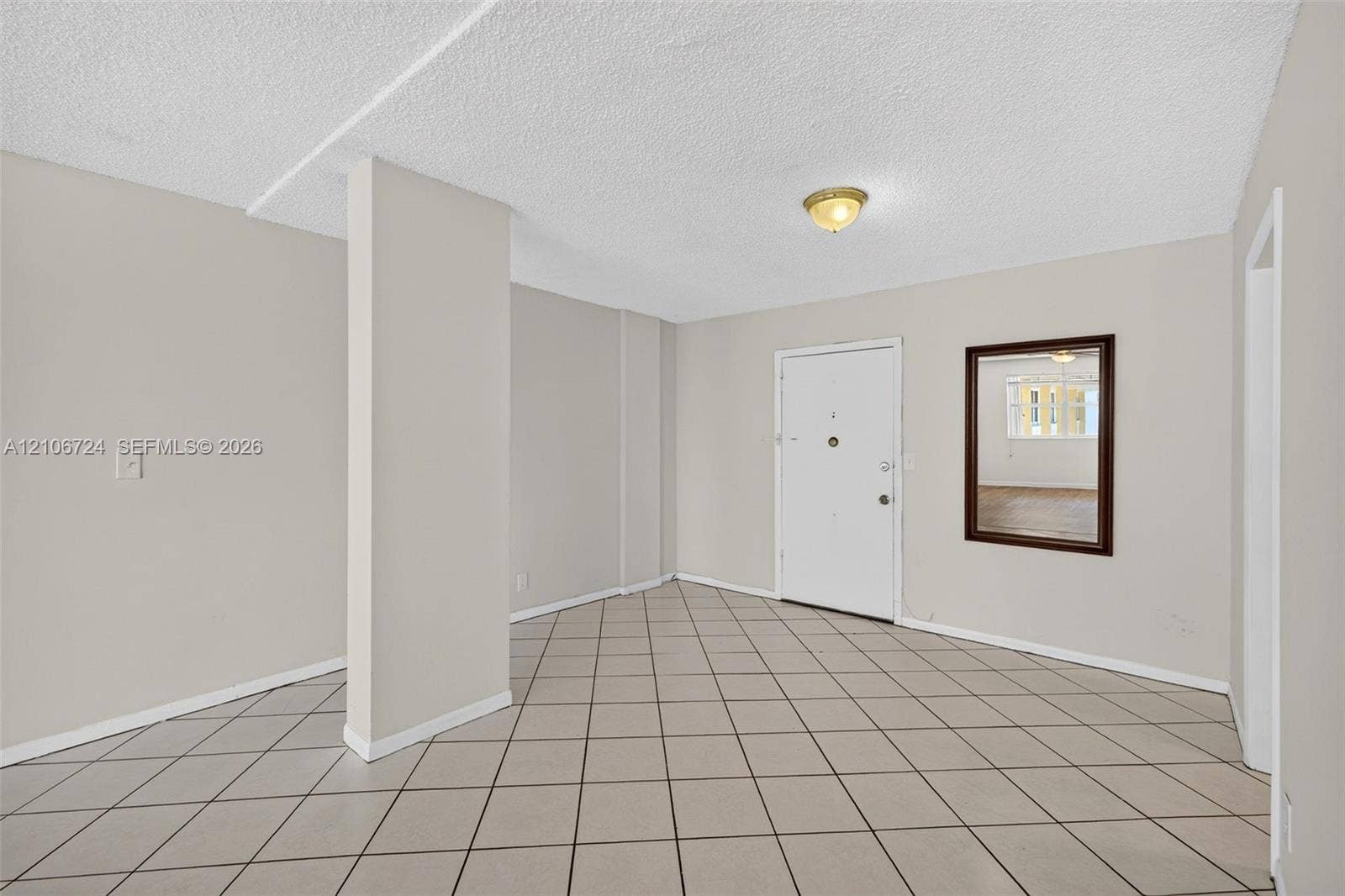 Neutral-toned interior space featuring ceramic tile flooring, a textured ceiling, a flush-mount light fixture, and a wall-mounted mirror